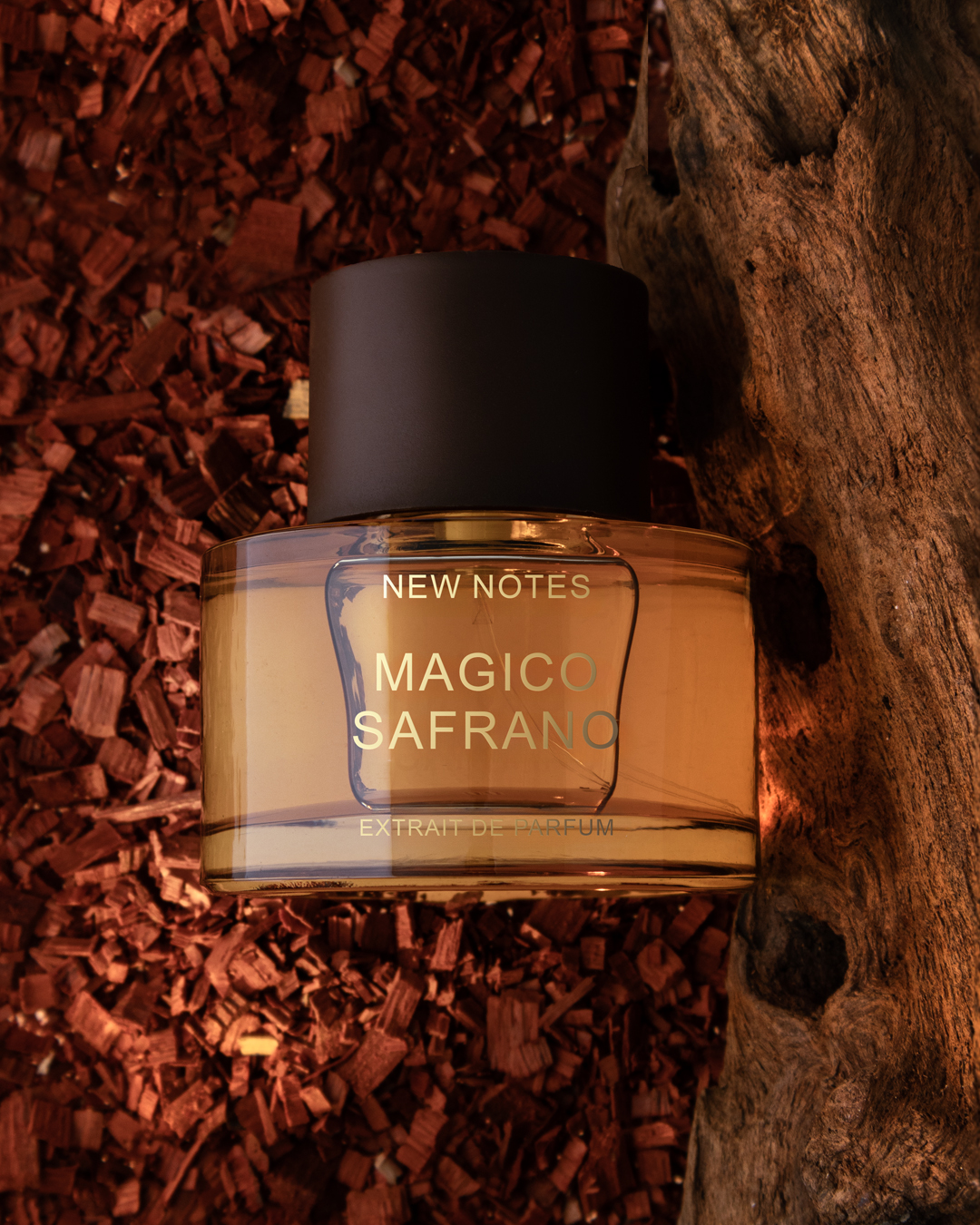 Flakon perfum New Notes Magico Safrano Extrait de Parfum na tle czerwonych wiórów drzewnych i naturalnego kawałka drewna. Ciepła, bursztynowa barwa flakonu podkreśla orientalny charakter zapachu inspirowanego szafranem i przyprawami.