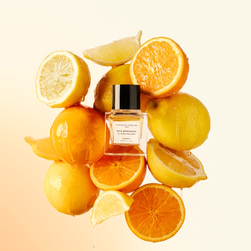 Flakon perfum Essential Parfums Nice Bergamote Extrait ustawiony w kompozycji z połówkami i ćwiartkami cytrusów — pomarańczy, cytryn i bergamotek. Sok z owoców spływa po butelce, podkreślając świeżość i soczystość zapachu. Tło w jasnych, ciepłych odcieniach kremu i pomarańczy potęguje letni, energetyczny charakter kompozycji