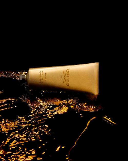 Złota, aluminiowa tuba kremu do rąk Creed Aventus Hand Cream leżąca na czarno-złotym tle przypominającym płynną teksturę lub odbłysk światła na powierzchni. Minimalistyczny, elegancki design opakowania z logo Creed podkreśla luksusowy charakter produktu.