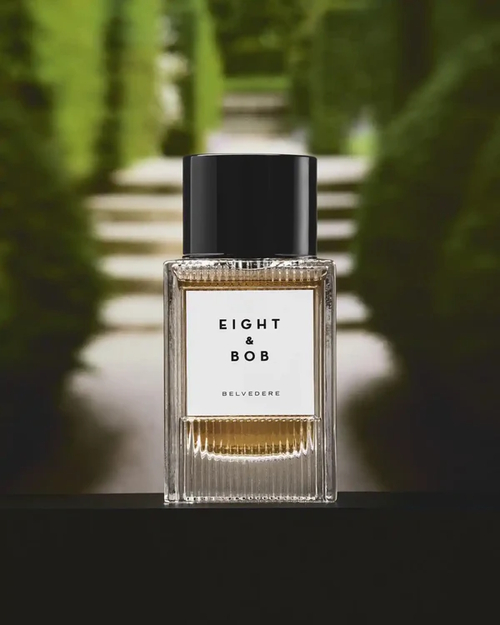 Flakon perfum Eight & Bob Belvedere w przezroczystym szkle z bursztynowym płynem i czarnym korkiem, ustawiony na tle eleganckiego ogrodu z zielonymi żywopłotami i schodami, tworząc atmosferę klasy i wyrafinowania.