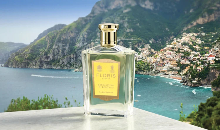 Na zdjęciu widoczny jest flakon perfum **Floris Bergamotto di Positano** ustawiony na jasnym kamiennym blacie. Elegancka, przezroczysta butelka z żółtą etykietą i złotym korkiem stoi na tle malowniczego wybrzeża Amalfi – skaliste góry schodzące do błękitnego morza, a na zboczu rozpościera się kolorowe miasteczko Positano. Obraz oddaje śródziemnomorski charakter zapachu – świeżość, elegancję i nostalgiczny urok włoskiego wybrzeża