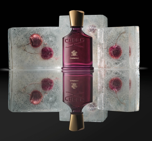 Flakon perfum Creed Carmina w odcieniu głębokiego burgundu z pozłacaną zatyczką, ustawiony na lustrzanej powierzchni w otoczeniu bloków lodu z zatopionymi czereśniami – elegancka kompozycja podkreślająca zmysłowy charakter zapachu.