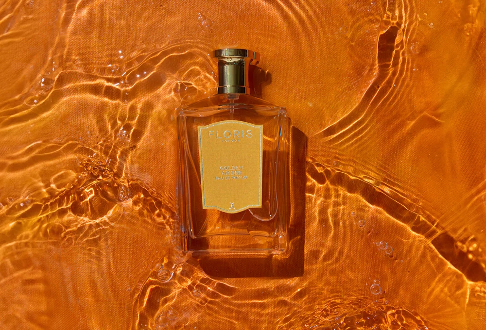 Flakon perfum floris golden amber leżący na pomarańczowym tle z efektem lśniącej wody i refleksami świetlnymi, które przywodzą na myśl ciepło słońca i bursztynowe odcienie zachodu słońca
