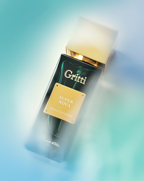 Na zdjęciu widoczny jest flakon perfum **Gritti Super Nova** z kolekcji Venetia. Buteleczka ma głęboki, ciemnozielony kolor z eleganckim złotym korkiem i złotą tabliczką z nazwą zapachu. Całość otulona jest miękką, niebiesko-białą mgłą, nadającą zdjęciu eteryczny, kosmiczny charakter. Flakon zdaje się unosić w przestrzeni, niczym tytułowa supernowa – pełna energii, światła i tajemnicy. Kompozycja wizualna podkreśla nowoczesność, intensywność i wyrafinowaną aurę zapachu.
