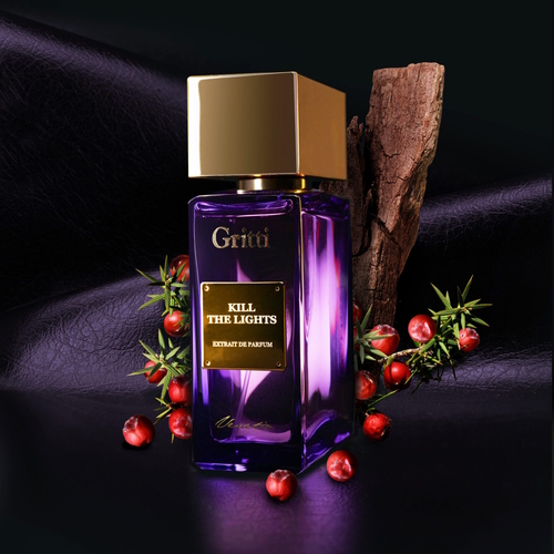 Na zdjęciu widoczny jest flakon perfum **Gritti Kill the Lights** z kolekcji *Venetia*. Buteleczka ma intensywnie fioletowy kolor, zwieńczona została eleganckim, złotym korkiem oraz złotą tabliczką z nazwą zapachu. Flakon ustawiono na tle czarnej skóry, w towarzystwie gałązek jałowca z czerwonymi jagodami oraz kawałka drewna, co symbolizuje głębię, intensywność i drzewno-aromatyczny charakter kompozycji. Całość emanuje tajemniczością, elegancją i nocnym klimatem zmysłowej prowokacji.