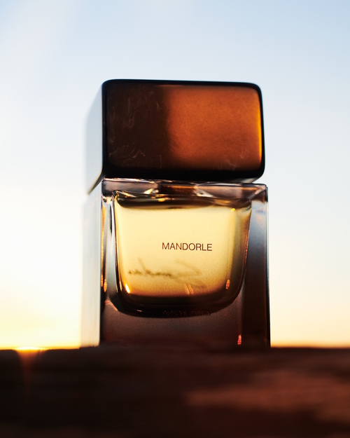 Zdjęcie flakonu perfum Sora Dora Mandorle sfotografowanego o zachodzie słońca. Butelka ma elegancki, minimalistyczny design – prostokątny kształt z grubego szkła, wypełniona złocistym płynem. Na środku widnieje napis „MANDORLE”. Czarne, masywne wieczko kontrastuje z ciepłym światłem tła, nadając całości luksusowego i zmysłowego charakteru.