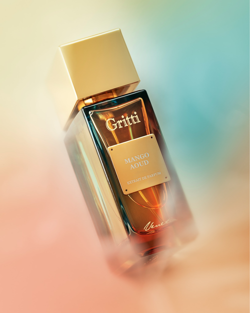 Na zdjęciu przedstawiono elegancki flakon perfum **Gritti Mango Aoud** z kolekcji *Venetia*. Butelka ma głęboki, bursztynowo-złoty odcień z połyskującą, złotą nakrętką oraz złotą tabliczką z nazwą zapachu. Całość spowita jest mglistym gradientem w ciepłych tonach mango, brzoskwini i chłodnego błękitu, co nadaje kompozycji zmysłowy, egzotyczny i nowoczesny charakter. Flakon wydaje się unosić w świetlnej poświacie, podkreślając luksusowe i orientalno-owocowe oblicze zapachu.