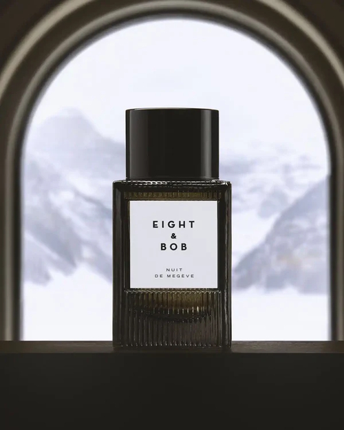 Flakon perfum Eight & Bob Nuit de Megève w ciemnym, eleganckim szkle z czarnym korkiem, ustawiony na tle ośnieżonych gór widocznych przez łukowate okno, tworząc luksusowy i zimowy klimat.