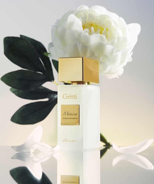 Flakon perfum Gritti Monica o matowym, białym wykończeniu i złotej zatyczce, ustawiony obok białej piwonii, symbolizującej główną nutę zapachu. Kompozycja zdjęcia utrzymana jest w eleganckim, minimalistycznym stylu