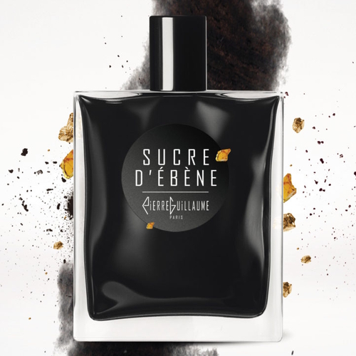 Flakon perfum Pierre Guillaume Sucre d’Ébène w eleganckiej, czarnej butelce z białym napisem na etykiecie. Za flakonem widać rozsypany brązowy cukier i ciemne tło przypominające kakao lub ziemię, co podkreśla słodko-drzewny charakter zapachu. Styl minimalistyczny, luksusowy i wyrafinowany