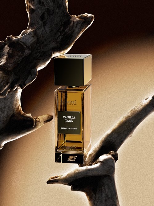 Flakon perfum Gritti Vanilla Tanà Extrait de Parfum o prostokątnym kształcie i złotej zatyczce stoi pomiędzy fragmentami drewna w ciepłym, złocisto-brązowym świetle. Elegancka, minimalistyczna butelka podkreśla luksusowy charakter zapachu, a otoczenie nawiązuje do naturalnych, drzewnych akordów kompozycji