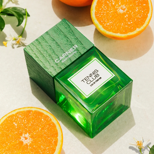 Zielony flakon perfum Carner Barcelona Tennis Club Eau de Parfum, ustawiony na jasnym tle w otoczeniu przekrojonych pomarańczy i drobnych białych kwiatów pomarańczy. Flakon ma elegancką, drewnianą nakrętkę w odcieniu zieleni i prostą etykietę z nazwą zapachu.
