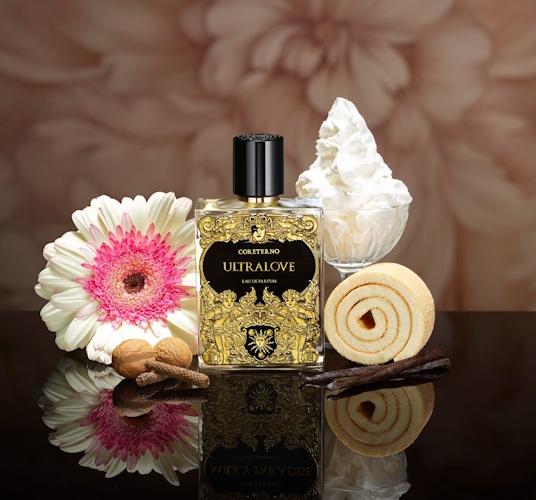Flakon perfum Coreterno Ultralove w złoto-czarnej, bogato zdobionej butelce stoi na lustrzanej powierzchni. Obok znajdują się elementy inspirowane nutami zapachu: kwiat gerbery, bita śmietana, orzechy laskowe, laska wanilii oraz zwinięte kruche ciasteczko. Całość utrzymana w ciepłej, kremowo-gourmandowej estetyce, podkreślającej słodki, zmysłowy charakter perfum