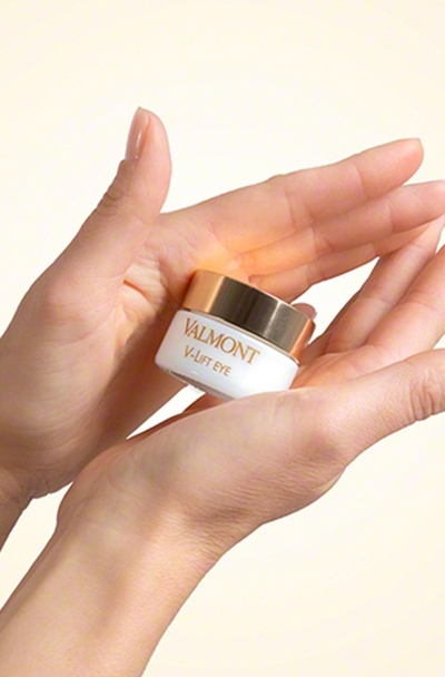 Słoiczek kremu pod oczy Valmont V-Lift Eye trzymany w dłoniach na jasnym, neutralnym tle. Eleganckie opakowanie z białym korpusem i złotą zakrętką podkreśla luksusowy charakter produktu i jego działanie liftingujące
